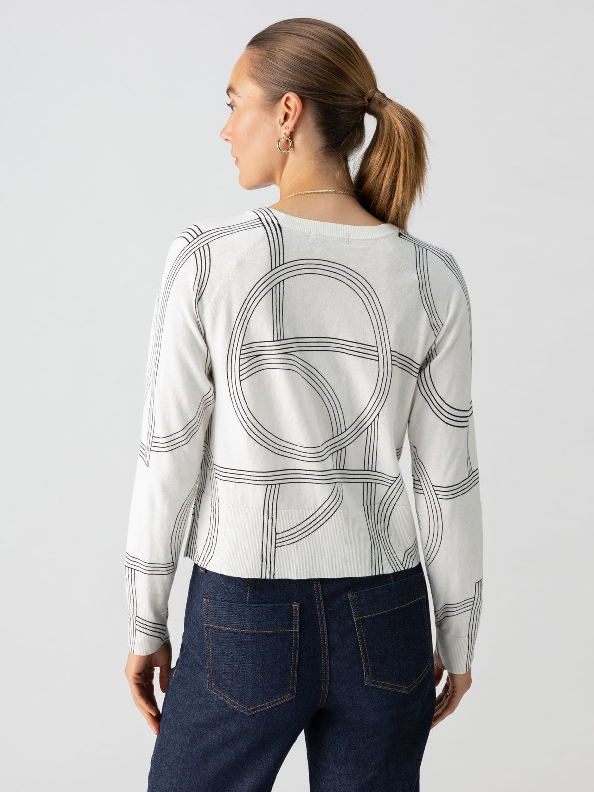 Seen from the back, a woman with her hair tied in a low ponytail wears the "All Day Long Sweater Graphic Lines" by Sanctuary Clothing. She pairs this white long-sleeve sweater, adorned with black circular patterns, with dark blue jeans and is accessorized with hooped earrings. The scene is set against a plain, light gray background.