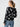 A person with long hair tied in a ponytail is seen from the back, wearing the All Day Long Sweater Rose Pop from Sanctuary Clothing, adorned with a white rose pattern. They are also wearing blue jeans and hoop earrings, standing against a plain light gray background.