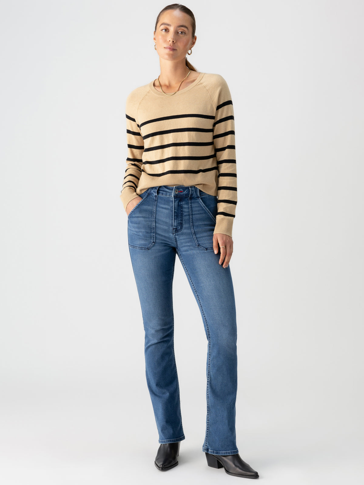 A person is standing against a plain background, wearing the "All Day Long Sweater True Khaki Black Stripe" by Sanctuary Clothing, blue high-waisted jeans, black ankle boots, and gold hoop earrings. They have their hands in their pockets and are looking at the camera.