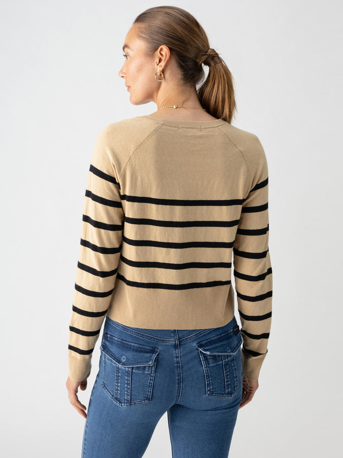 A woman with brown hair pulled into a ponytail is seen from the back, wearing Sanctuary Clothing's All Day Long Sweater in True Khaki Black Stripe paired with blue jeans. She accessorizes with gold hoop earrings and a necklace, set against a light grey background.