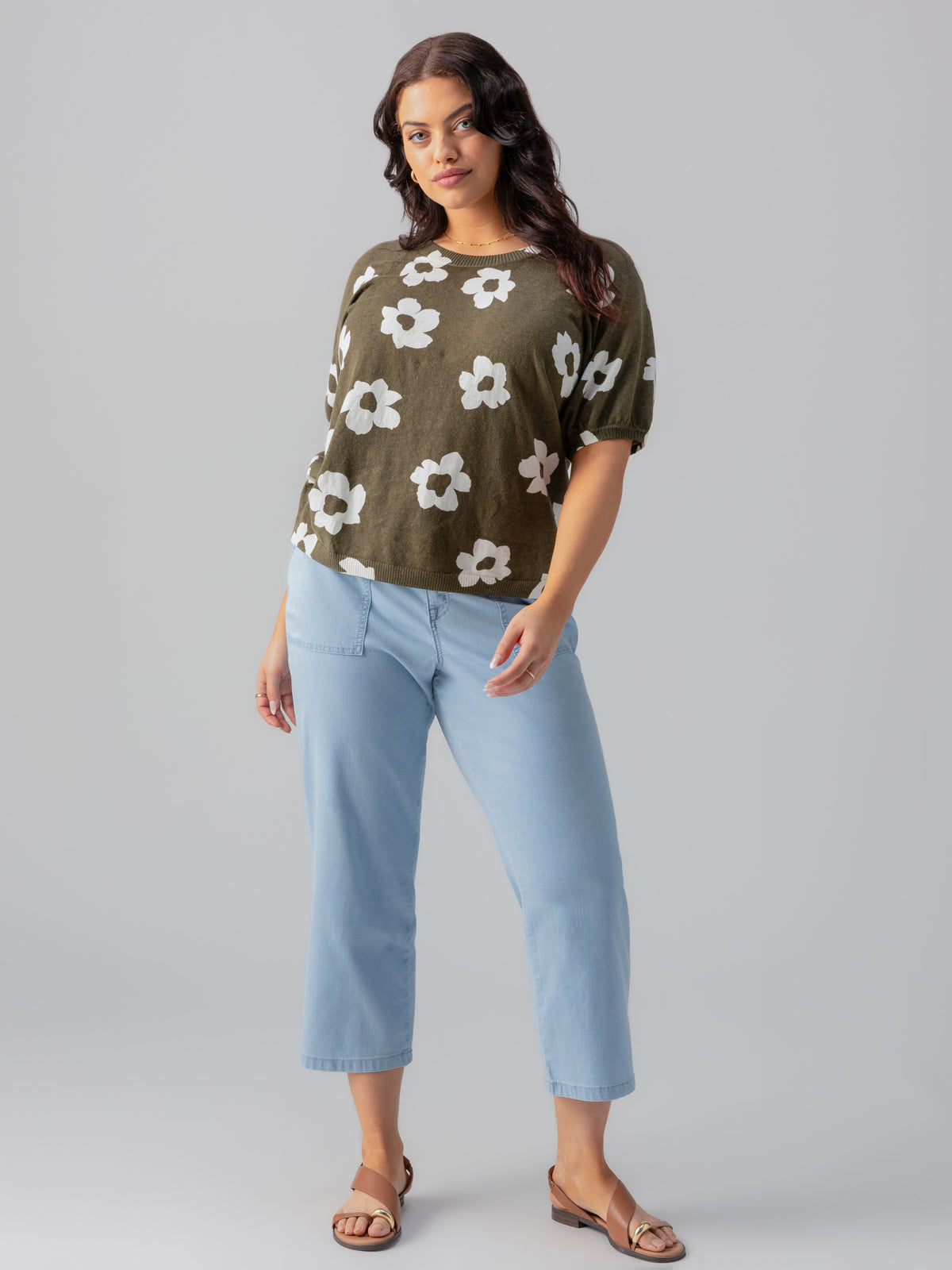 A person with long dark hair stands against a gray background. They are wearing the Sanctuary Clothing "Sunny Days Sweater" in Burnt Olive from the Pop Inclusive Collection, light blue cropped jeans, and brown sandals. They have a relaxed posture with one hand resting on their hip.