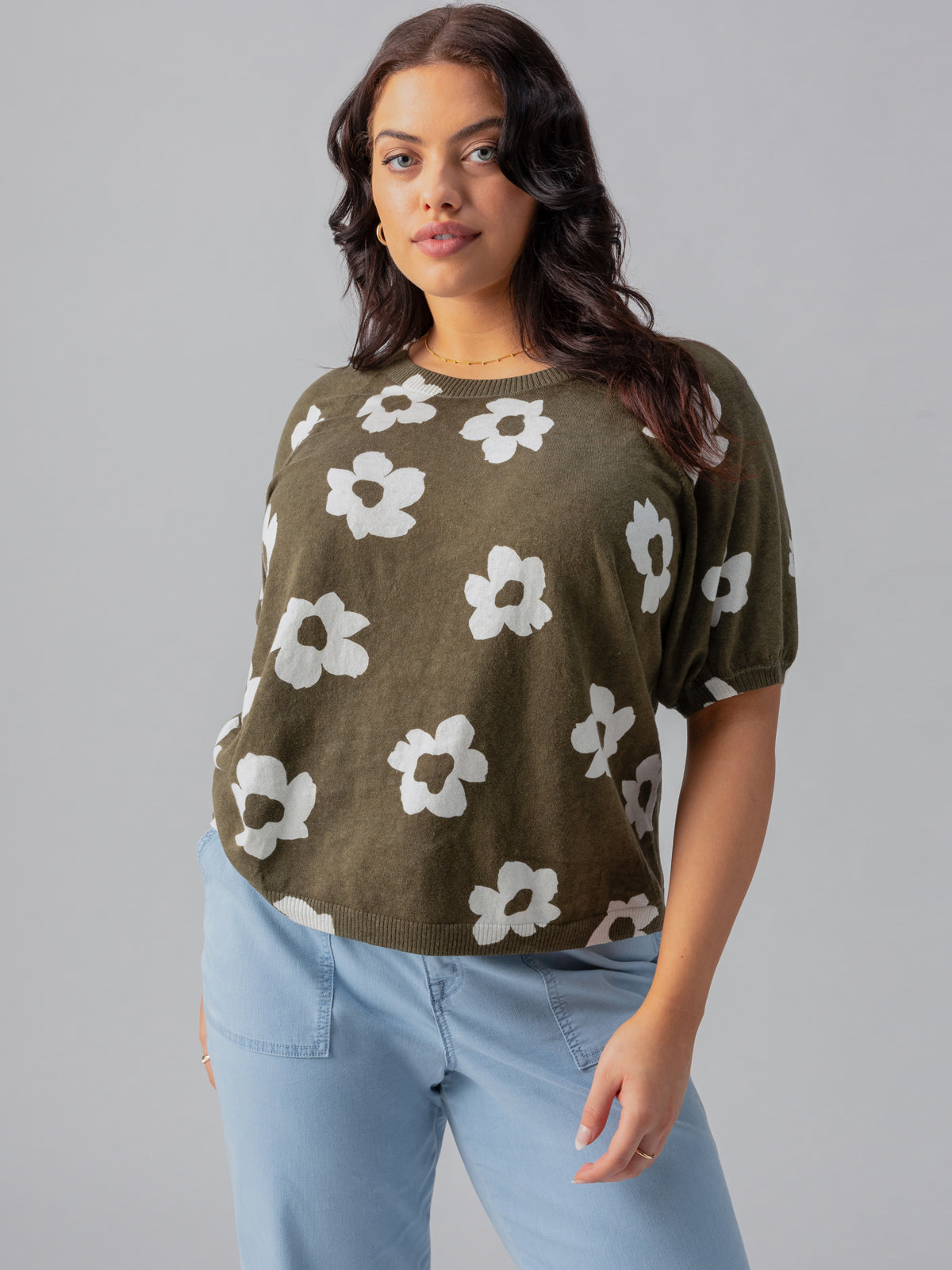 A woman stands confidently against a gray backdrop wearing the Sunny Days Sweater Burnt Olive Pop from Sanctuary Clothing's Inclusive Collection, featuring a relaxed fit and a white floral pattern. She pairs it with light blue pants, has long wavy hair, and accessorizes with small hoop earrings. Her hands are in her pockets.