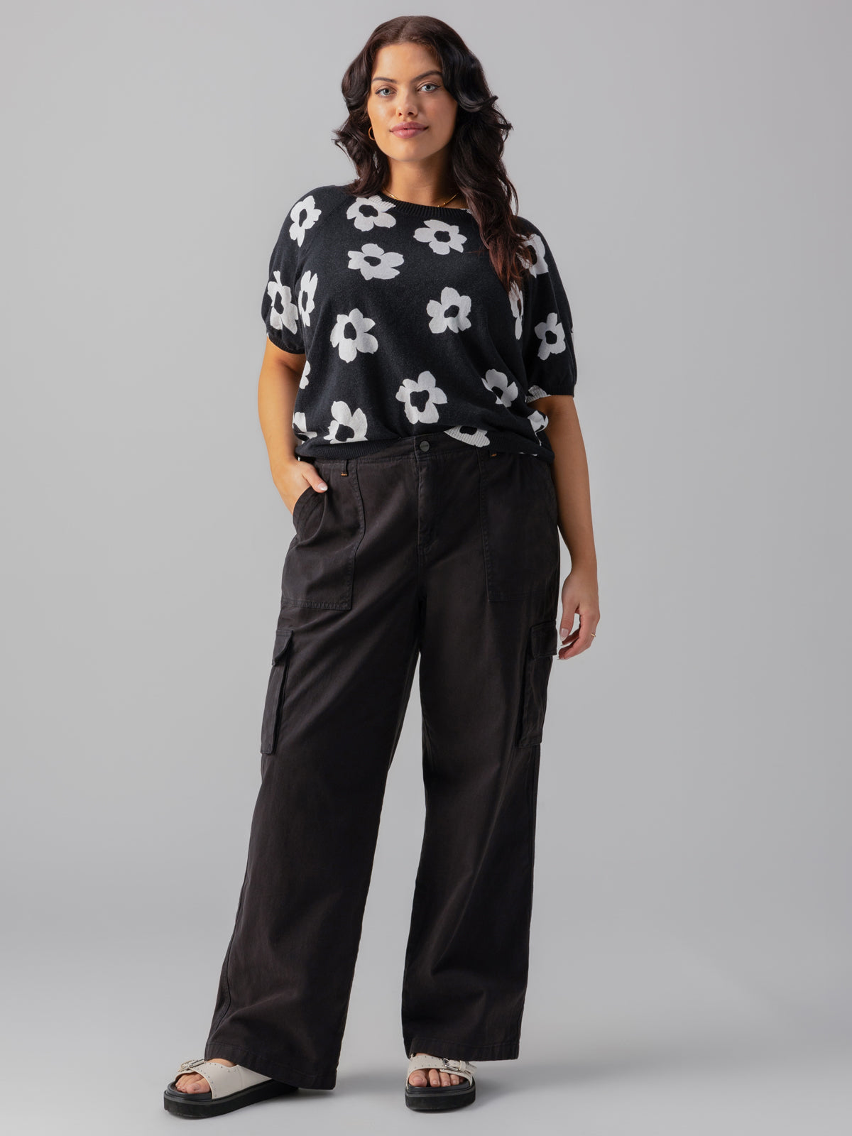 A person with long, wavy hair stands confidently against a gray background. They are wearing the Sunny Days Sweater Flower Pop from Sanctuary Clothing's Inclusive Collection, black high-waisted cargo pants, and white sandals. Their hands are casually placed in their pants pockets, and they are smiling slightly.