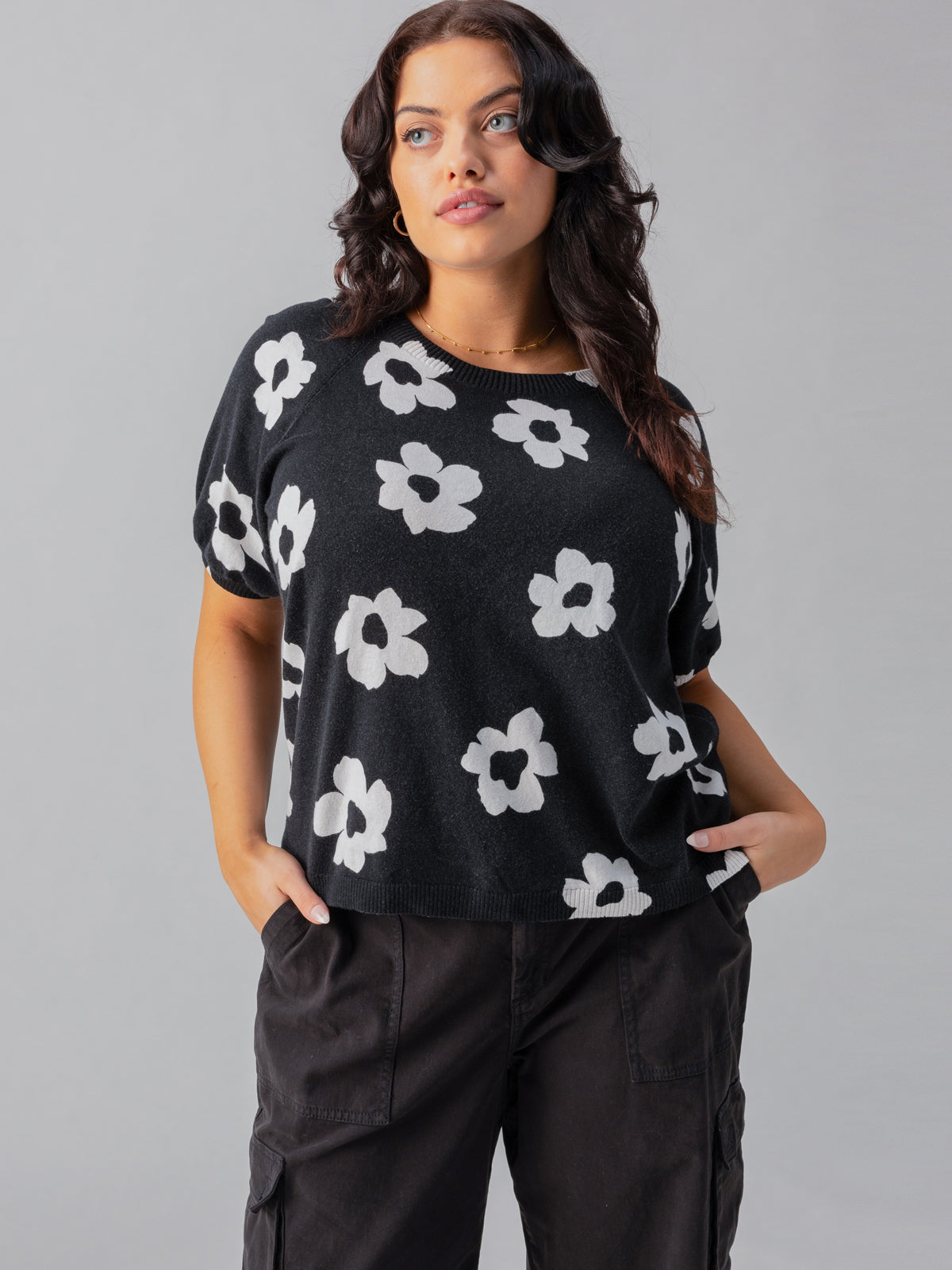 A woman with long, dark hair models the Sunny Days Sweater Flower Pop from Sanctuary Clothing's Inclusive Collection, paired with dark cargo pants and standing against a plain gray background, her hands tucked casually in her pockets.