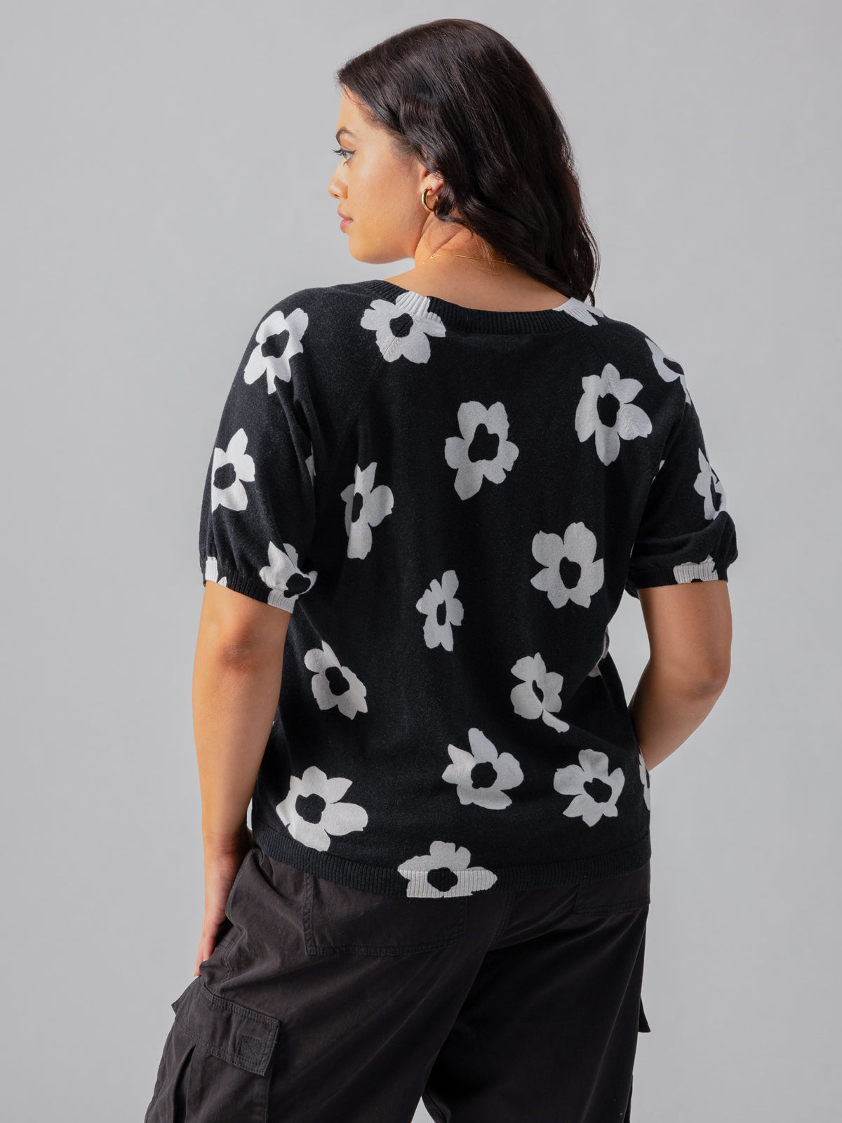 A person with shoulder-length dark hair is seen from behind, wearing Sanctuary Clothing's Sunny Days Sweater Flower Pop Inclusive Collection paired with black cargo pants. The background is a plain light gray.