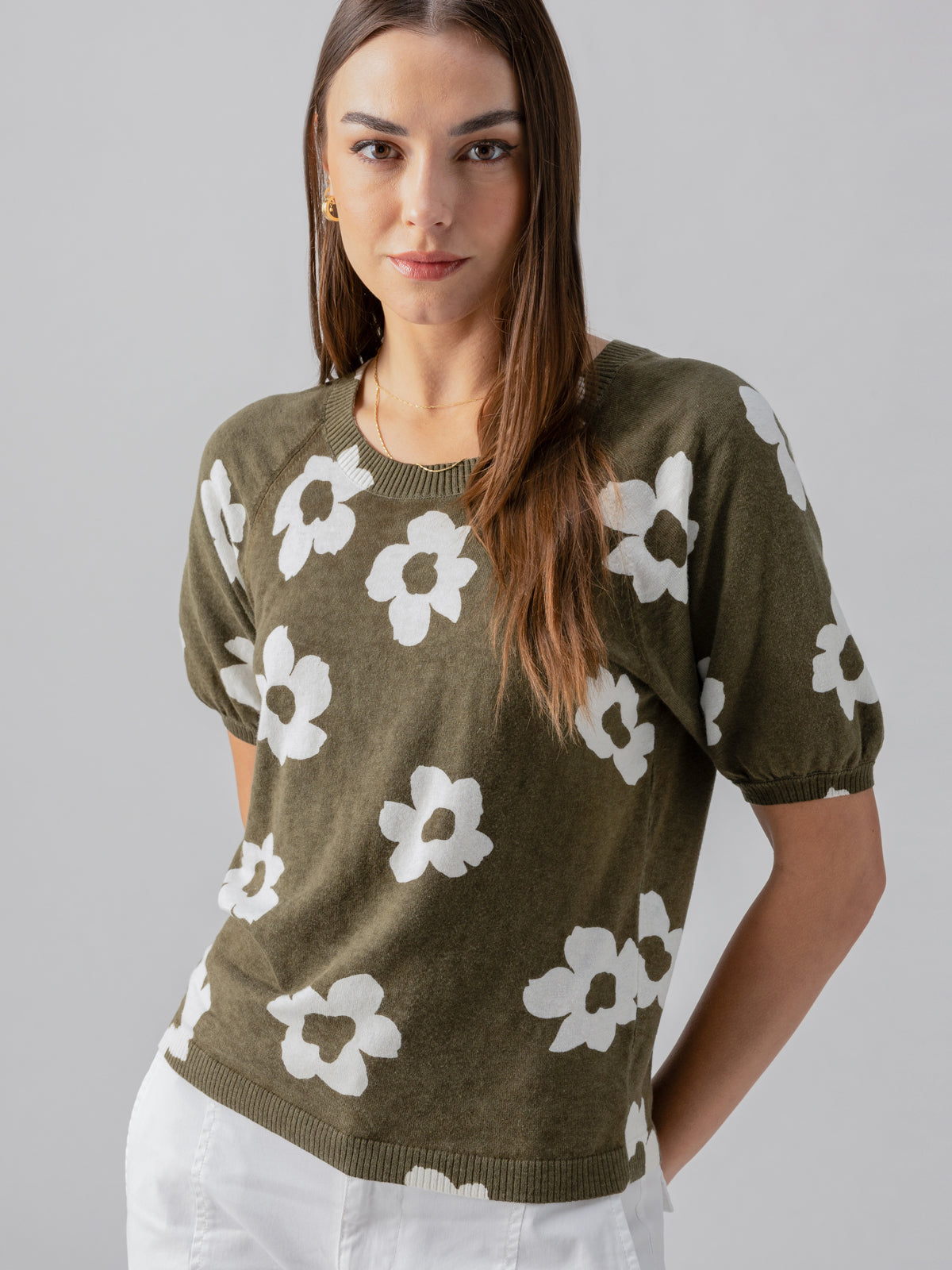 A woman with long brown hair is wearing the Sunny Days Sweater Burnt Olive Pop from Sanctuary Clothing, which is a green short-sleeved sweater with white flower patterns, paired with white pants. She is standing against a plain background, smiling slightly with her hands behind her back.