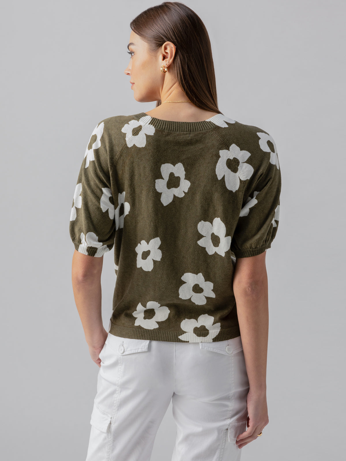 A woman with long hair is turned away from the camera, showcasing her Sunny Days Sweater Burnt Olive Pop by Sanctuary Clothing, adorned with white flower patterns. She is also wearing white pants. The background is plain light grey, emphasizing her outfit.