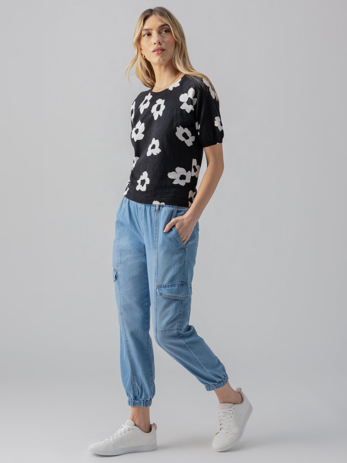 A woman stands against a gray background, wearing Sanctuary Clothing's Sunny Days Sweater Flower Pop—a black short-sleeve top with white flower prints—paired with light blue jogger pants featuring side pockets and white sneakers. Her hands rest in the pockets of her joggers as she gazes slightly to her left.