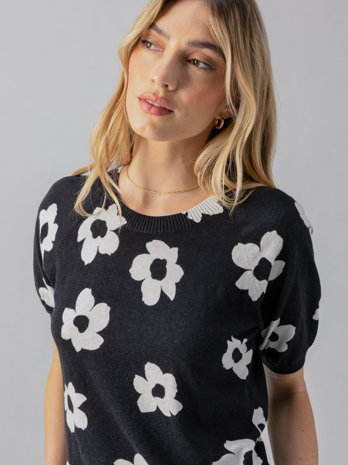 A person with shoulder-length blonde hair is wearing the Sanctuary Clothing Sunny Days Sweater Flower Pop, which features a black base adorned with white floral patterns. They have gold earrings and are gazing slightly downward, posing against a plain gray background.