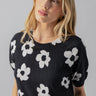 A person with shoulder-length blonde hair is wearing the Sanctuary Clothing Sunny Days Sweater Flower Pop, which features a black base adorned with white floral patterns. They have gold earrings and are gazing slightly downward, posing against a plain gray background.