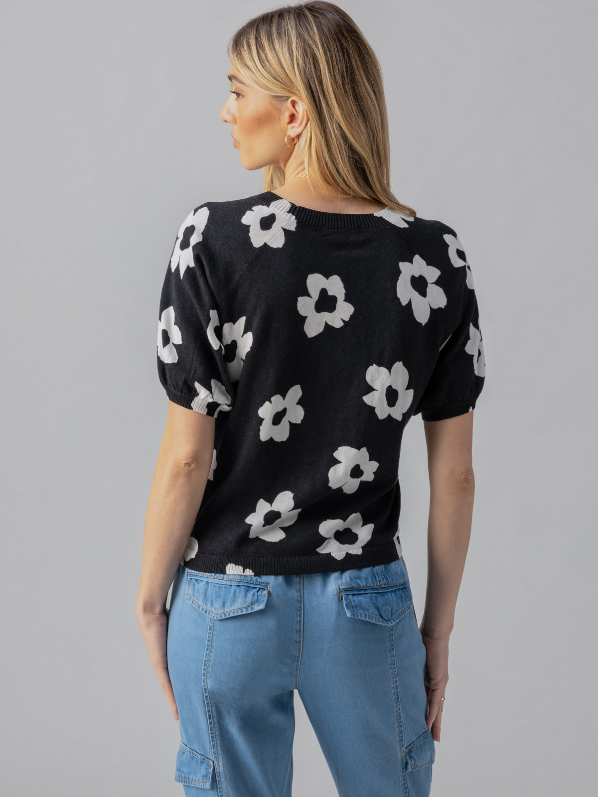 A woman with blonde hair is turned partially away from the camera, wearing the Sunny Days Sweater Flower Pop from Sanctuary Clothing, a black short-sleeve sweater adorned with large white flower patterns. She pairs it with light blue jeans that have side pockets. The background is plain gray.