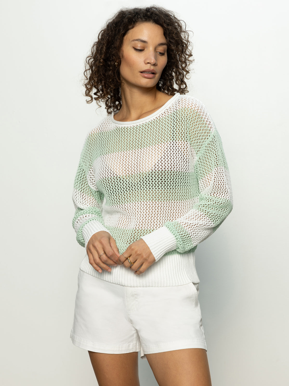 A person with curly hair stands against a light background, wearing Sanctuary Clothing's open knit sweater in mint tea chalk over a white top, paired with white shorts. They have their hands clasped lightly.