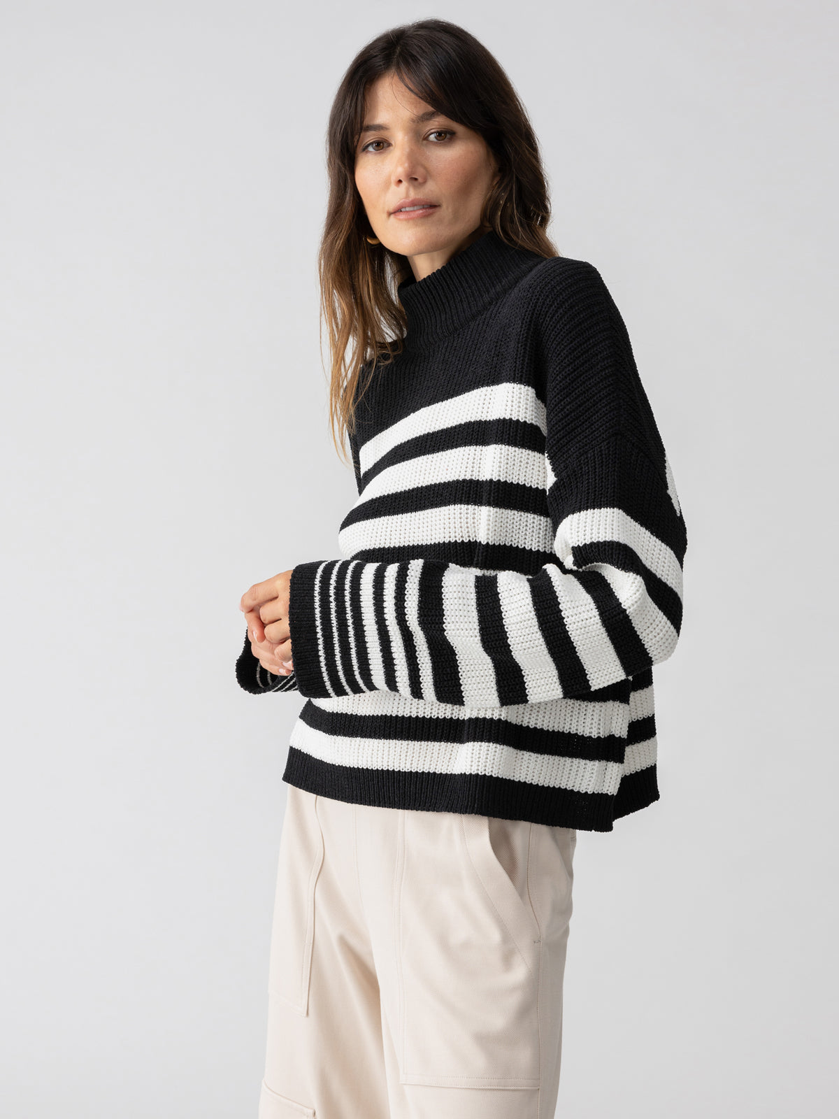 A person with long dark hair wears Sanctuary Clothing's Stay Cozy Sweater in Black Chalk Stripe paired with cream-colored pants. They are standing against a plain gray background, gazing slightly off-camera, with their hands casually clasped in front.