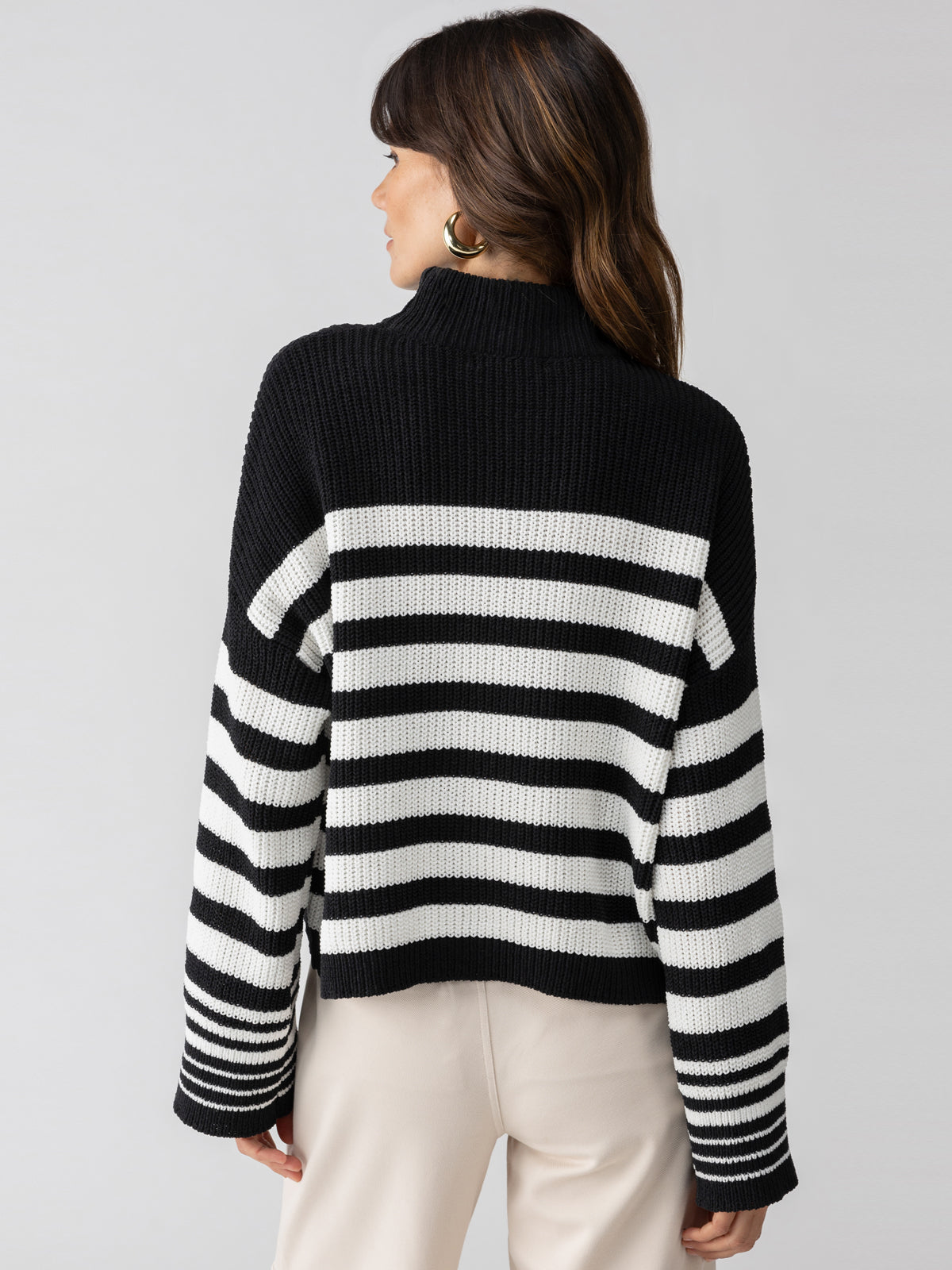 A woman with brown hair stands facing away, highlighting the Sanctuary Clothing Stay Cozy Sweater Black Chalk Stripe, featuring a high neckline and long sleeves. She pairs it with cream-colored pants against a plain gray background.