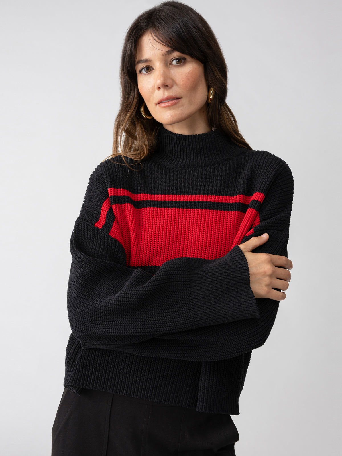 A person with long dark hair wearing the Sanctuary Clothing Stay Cozy Sweater Black Red Stripe stands against a plain gray background. Their hands are folded in front of them, and they have a neutral expression.