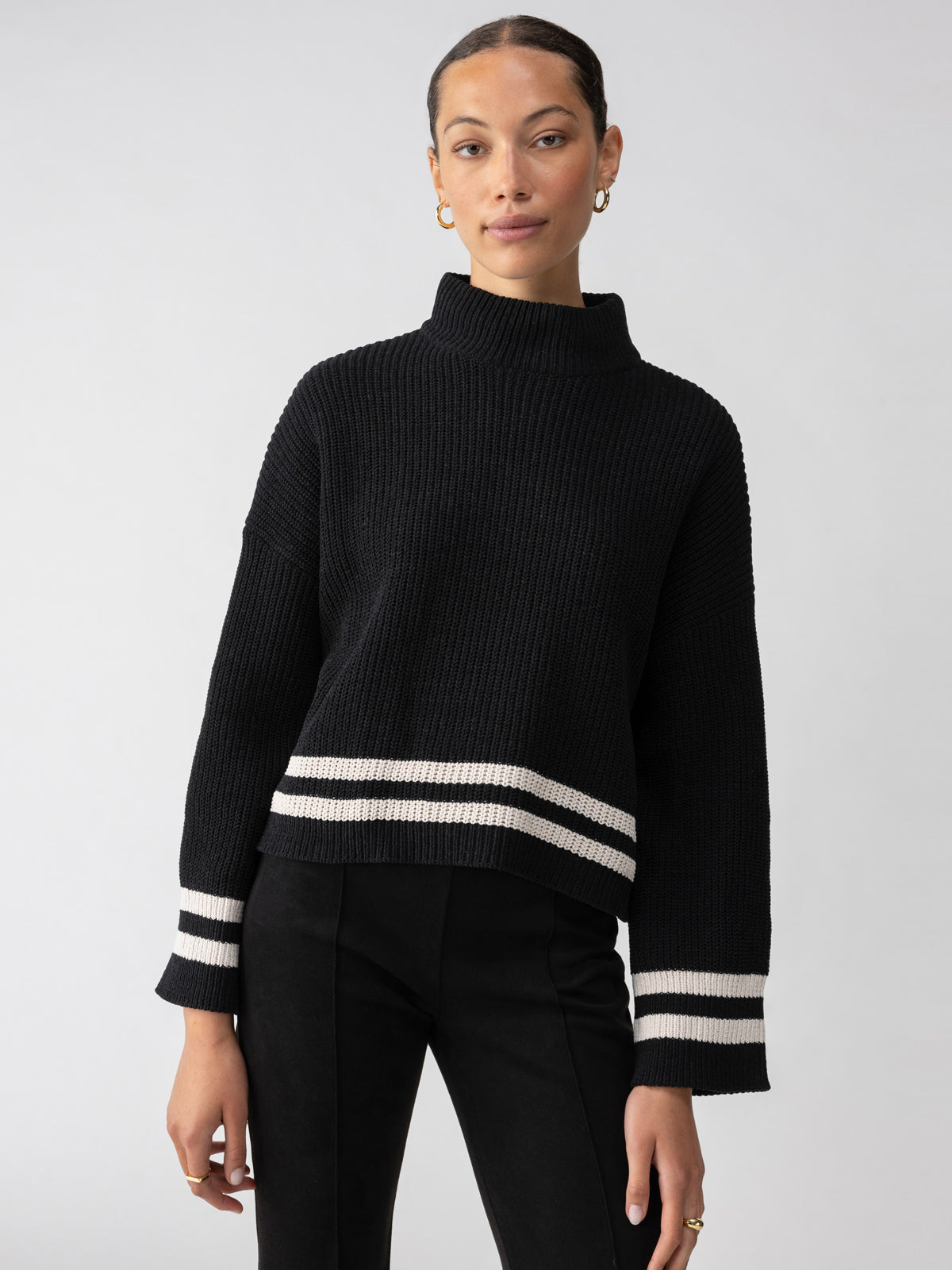 A person with a neat hairstyle is wearing the Stay Cozy Sweater Black Toasted Stripe by Sanctuary Clothing, paired with black pants. The sweater features white stripes near the cuffs and hem. The individual is accessorized with gold hoop earrings and positioned against a plain light-gray background.