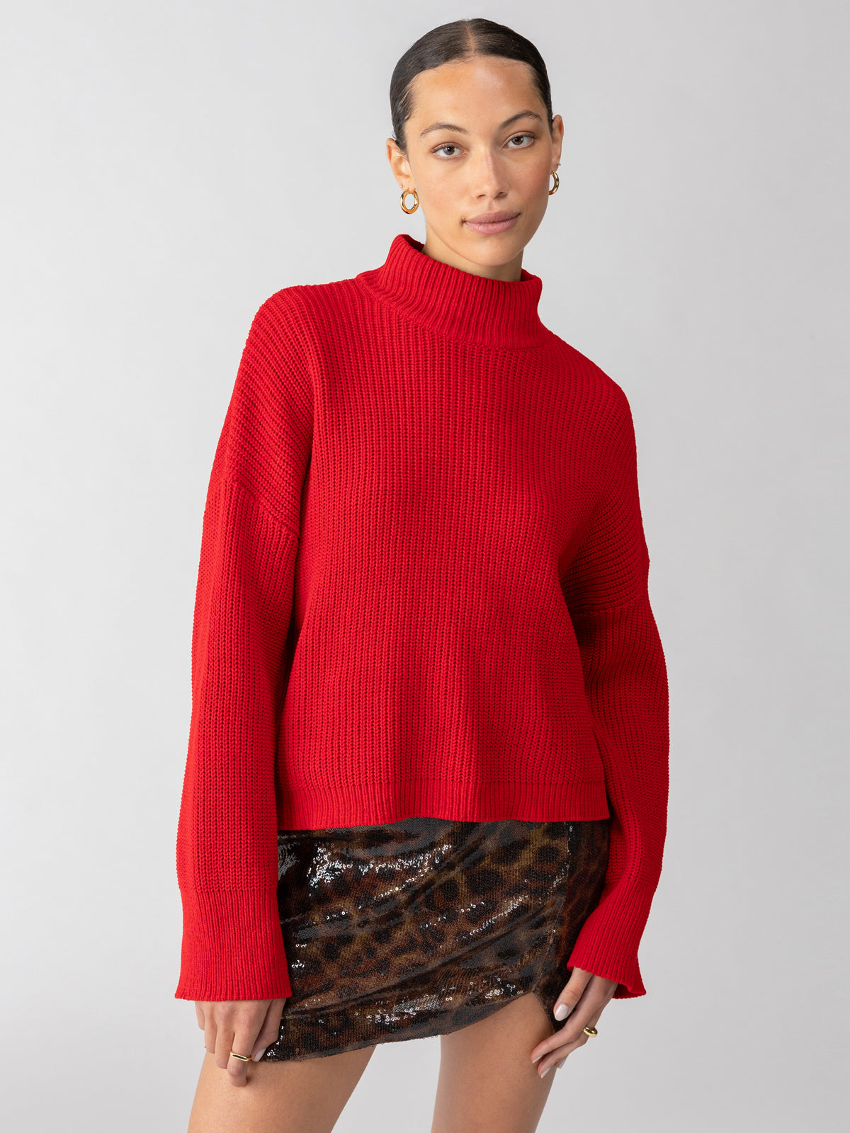 A person with a neat updo is wearing the Stay Cozy Sweater in Mars Red by Sanctuary Clothing, paired with a sequined leopard print mini skirt. The individual is also accessorizing with gold hoop earrings, standing against a plain light gray background.