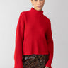 A person with a neat updo is wearing the Stay Cozy Sweater in Mars Red by Sanctuary Clothing, paired with a sequined leopard print mini skirt. The individual is also accessorizing with gold hoop earrings, standing against a plain light gray background.