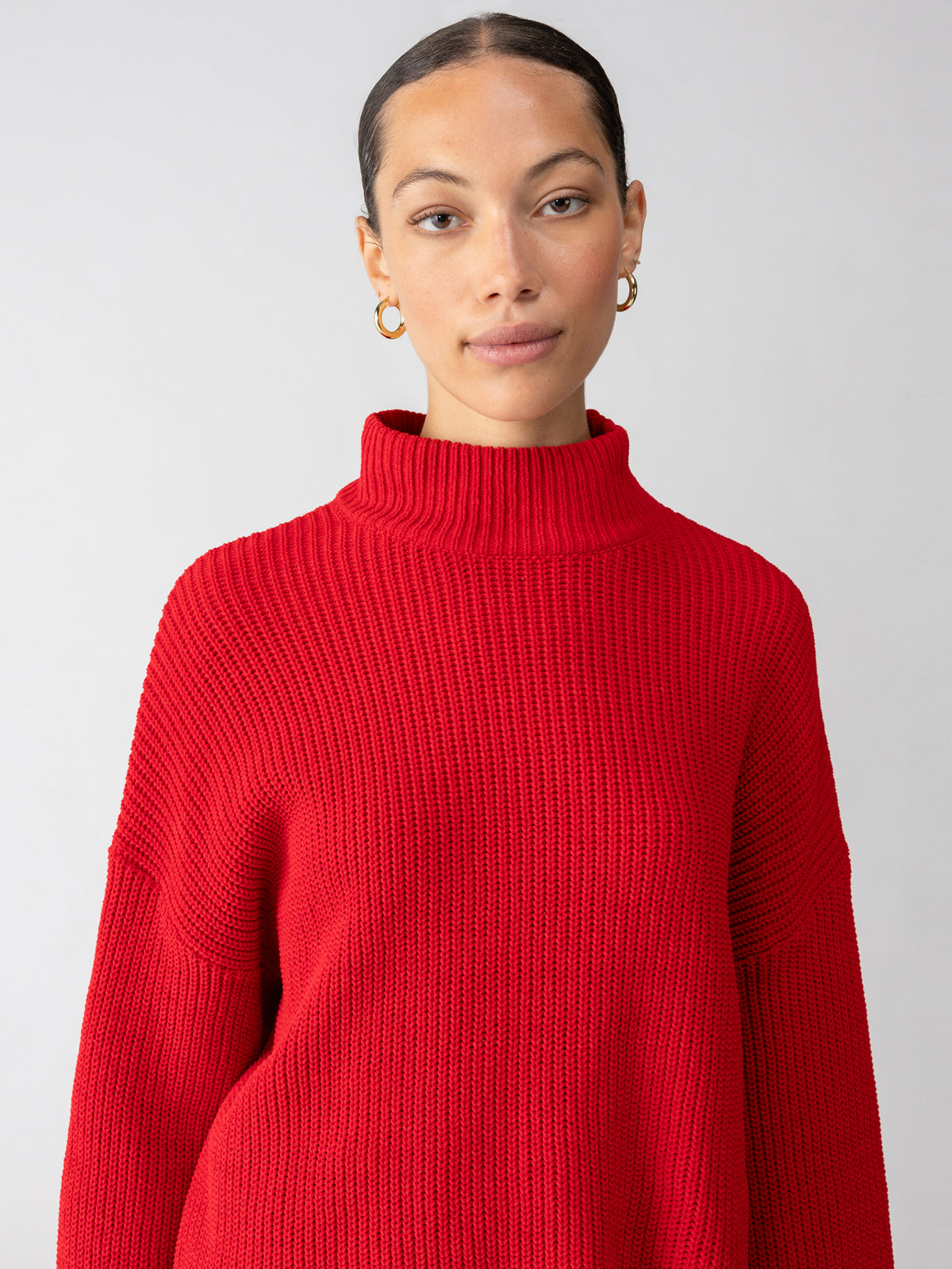 A person with pulled-back hair is wearing gold hoop earrings and a bright red Stay Cozy Sweater Mars Red from Sanctuary Clothing. The background is plain and light-colored, drawing attention to the individual's attire and face.