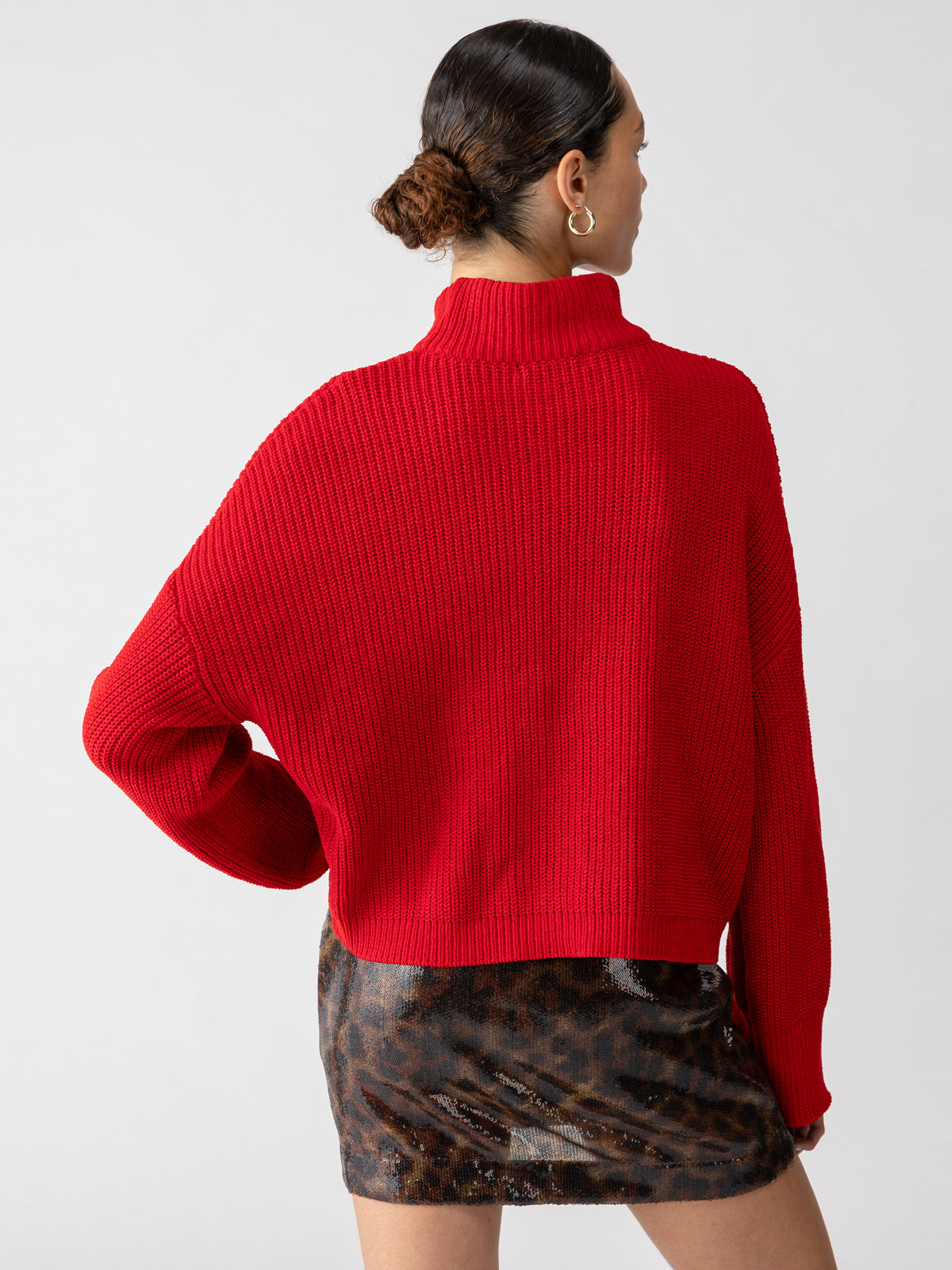 A person with their hair styled in a bun is wearing the Stay Cozy Sweater in Mars Red by Sanctuary Clothing, along with an animal print skirt. The photo is taken from behind, highlighting the back of the sweater and the intricate patterns on the skirt. The background is plain and light-colored.