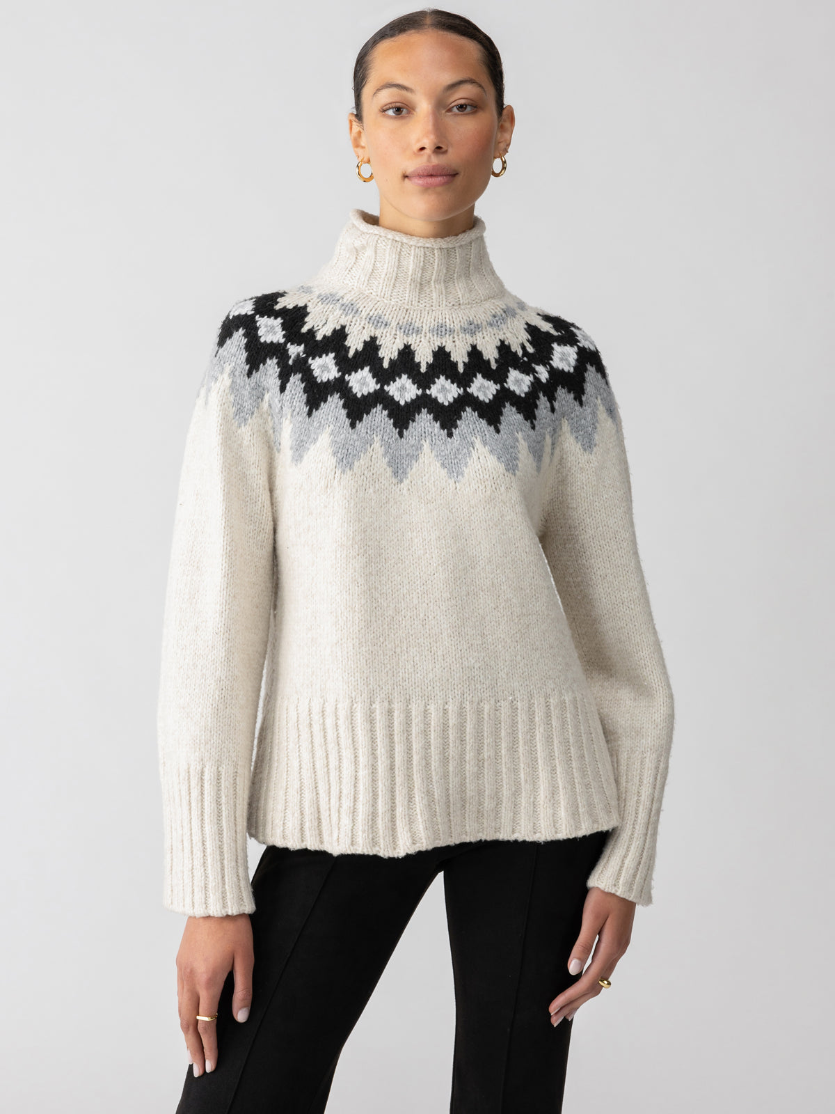 A person with tied-back hair and oval-shaped earrings wears the "Tis The Season Fairisle Sweater Toasted Almond Multi" by Sanctuary Clothing, a cream-colored knit with a high neck that showcases a black and grey zig-zag pattern across the chest. They pose against a plain white background while wearing black pants.