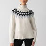 A person with tied-back hair and oval-shaped earrings wears the "Tis The Season Fairisle Sweater Toasted Almond Multi" by Sanctuary Clothing, a cream-colored knit with a high neck that showcases a black and grey zig-zag pattern across the chest. They pose against a plain white background while wearing black pants.