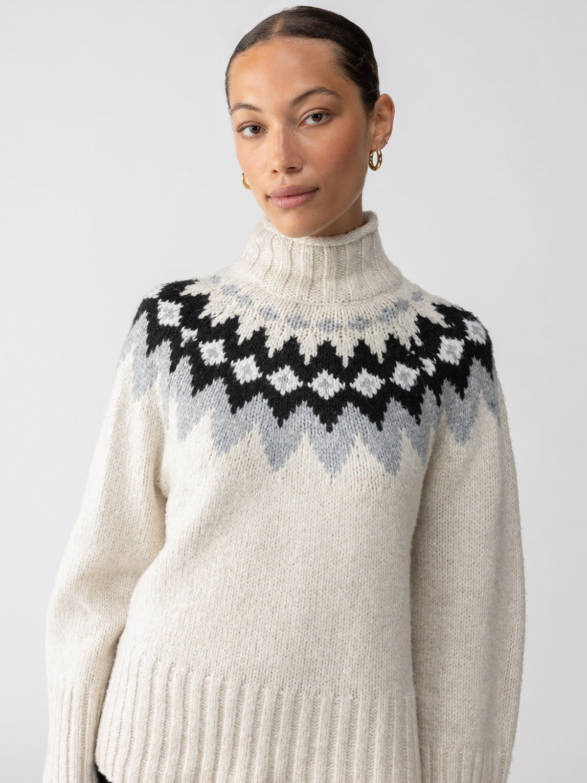 A person is seen wearing the Tis The Season Fairisle Sweater in Toasted Almond Multi by Sanctuary Clothing, featuring a geometric black, gray, and white pattern across the chest and shoulders. Their hair is pulled back and they are accessorized with gold hoop earrings, posing against a light gray background.