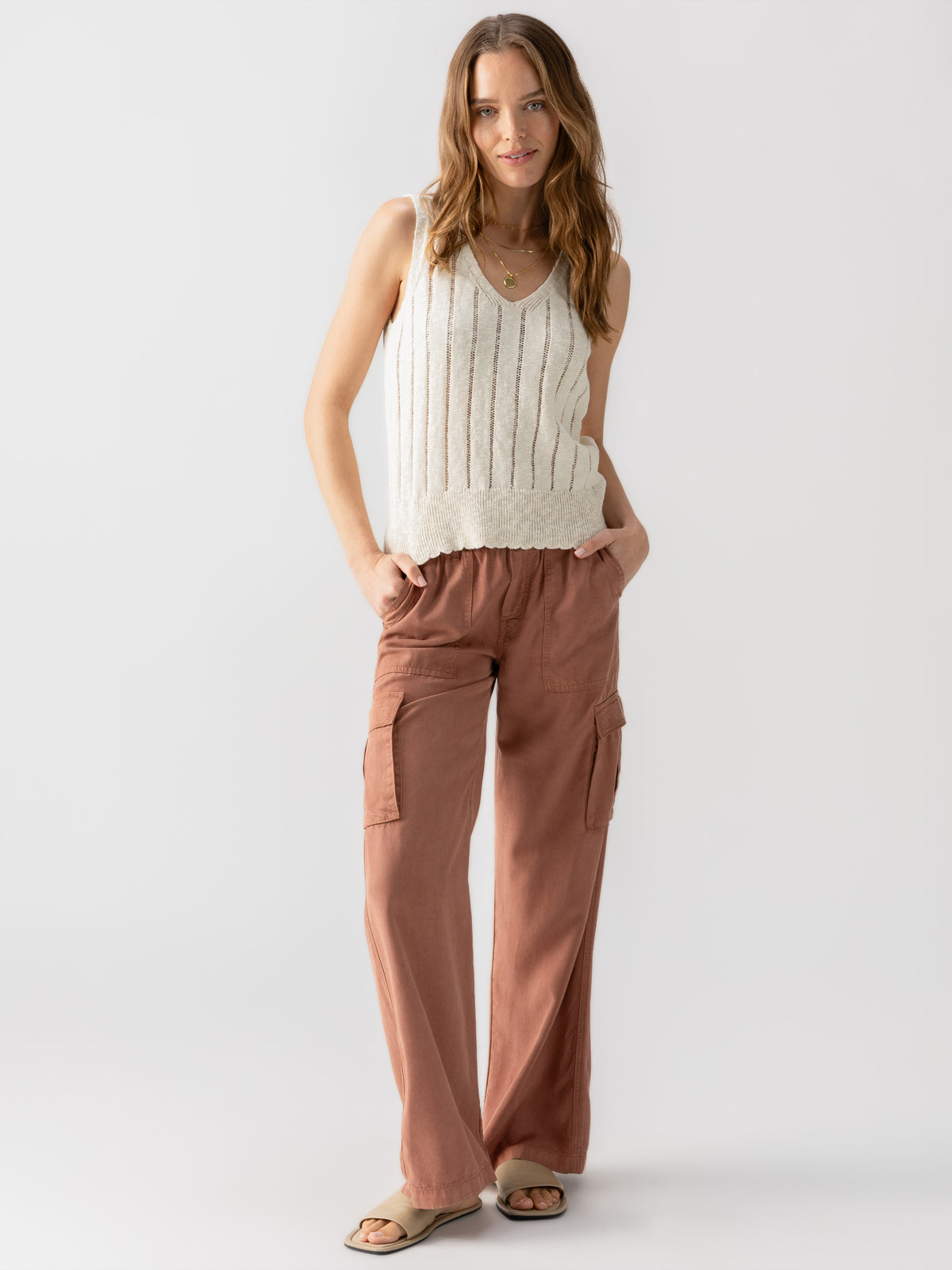 A woman with long, wavy hair stands against a plain white background. She is wearing the Sanctuary Clothing Vacation Mode Shell Birch, a sleeveless cream-colored knit top, and loose rust-colored cargo pants with large pockets. She has her hands in her pockets and is smiling gently while wearing sandals.