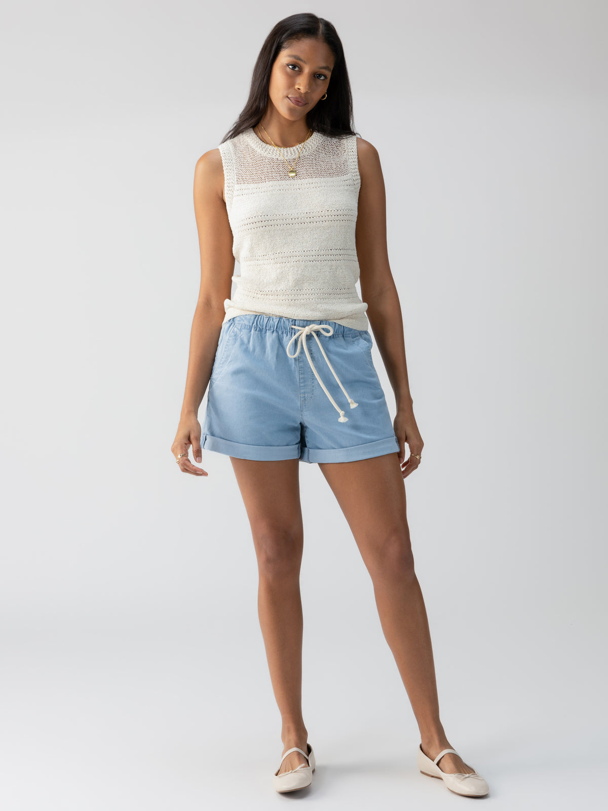 A woman with long dark hair is wearing the "Waves Of Summer Shell Birch" sleeveless white knit top by Sanctuary Clothing and light blue drawstring shorts. She has light-colored slip-on shoes and is standing against a plain light grey background, posing casually with her arms by her sides.