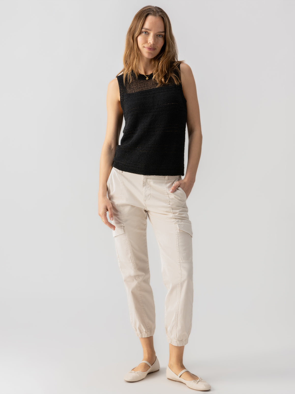 A woman with long hair stands against a plain background, wearing the 'Waves Of Summer Shell Black' top by Sanctuary Clothing. She is paired with light-colored pants featuring pockets and beige shoes. With one hand in her pocket and the other resting by her side, she gazes into the camera with a relaxed posture.