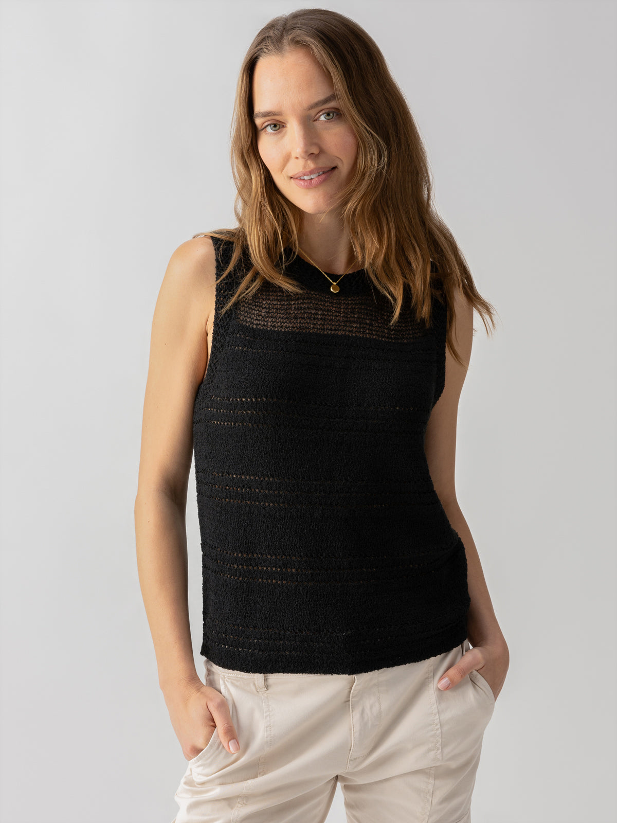 A person with long brown hair is wearing the Waves Of Summer Shell Black sleeveless top by Sanctuary Clothing and beige pants. They are standing against a plain background, smiling with their hands in their pockets. They are also wearing a small, round gold necklace.