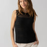 A person with long brown hair is wearing the Waves Of Summer Shell Black sleeveless top by Sanctuary Clothing and beige pants. They are standing against a plain background, smiling with their hands in their pockets. They are also wearing a small, round gold necklace.