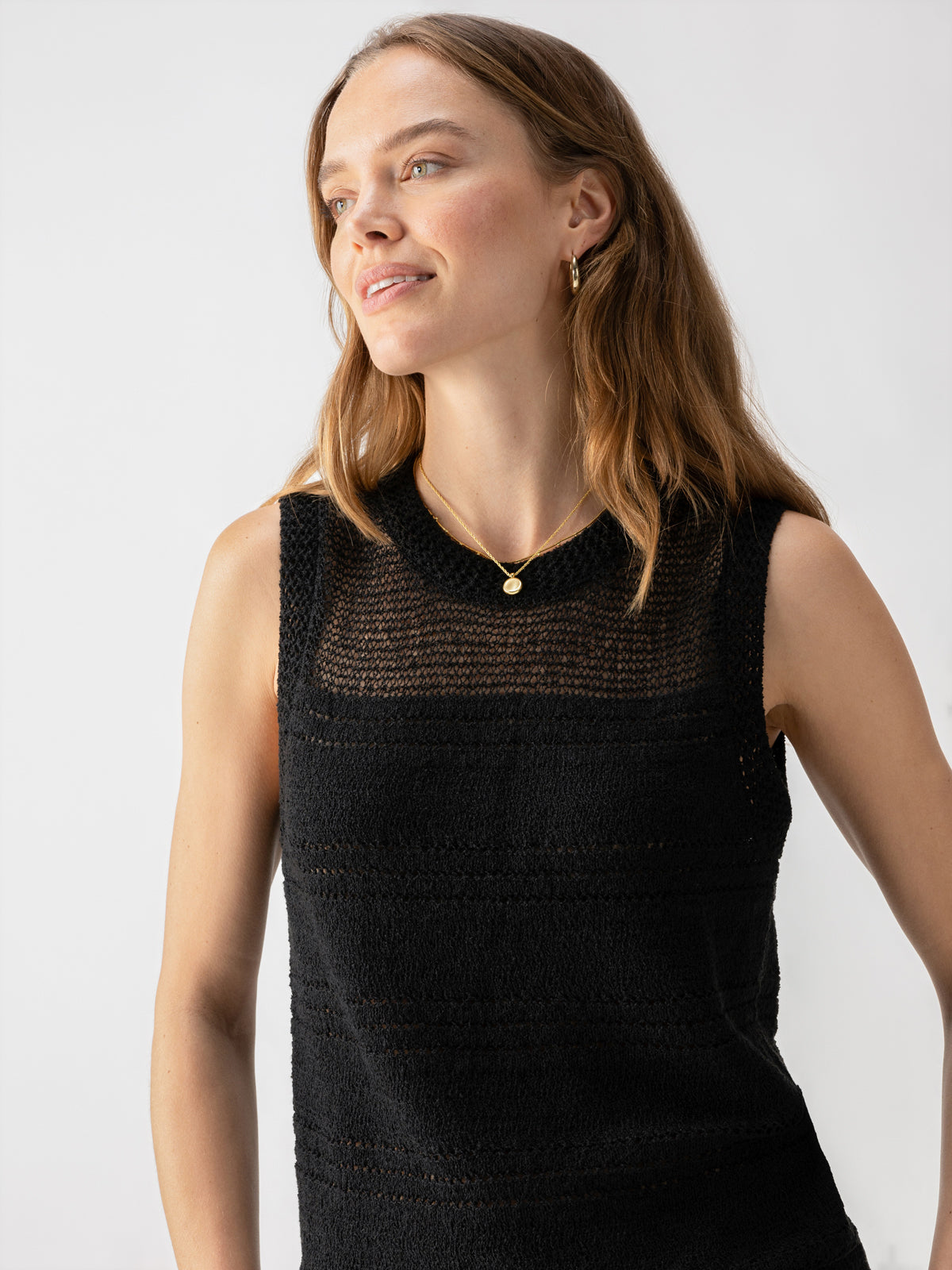 A woman with long, light brown hair smiles slightly while looking to her left. She is wearing the Waves Of Summer Shell Black sleeveless top by Sanctuary Clothing, along with gold hoop earrings and a matching necklace. The background is plain white.