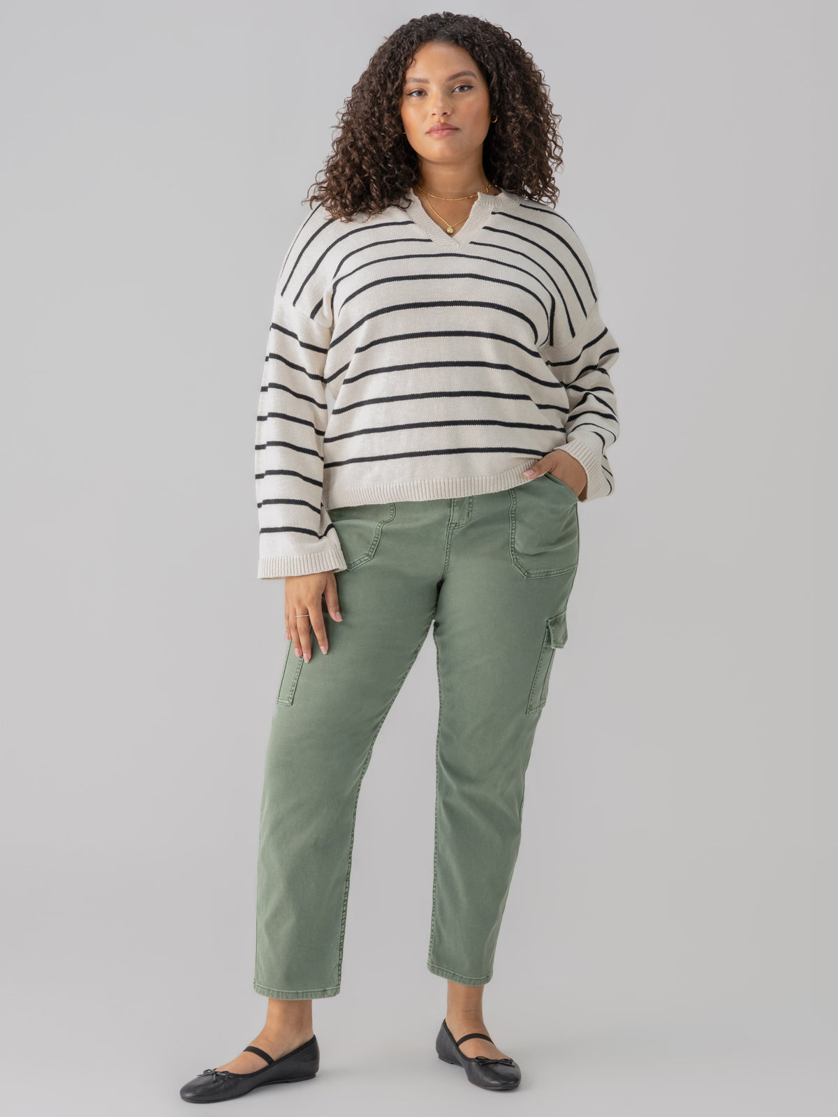 A woman with curly hair is wearing a Chill Vibes Sweater in Chalk Black Stripe from Sanctuary Clothing's Inclusive Collection, paired with green cargo pants. She stands against a plain gray background, posed with one hand in her pocket and the other relaxed by her side. She is wearing black flats and looking straight ahead.