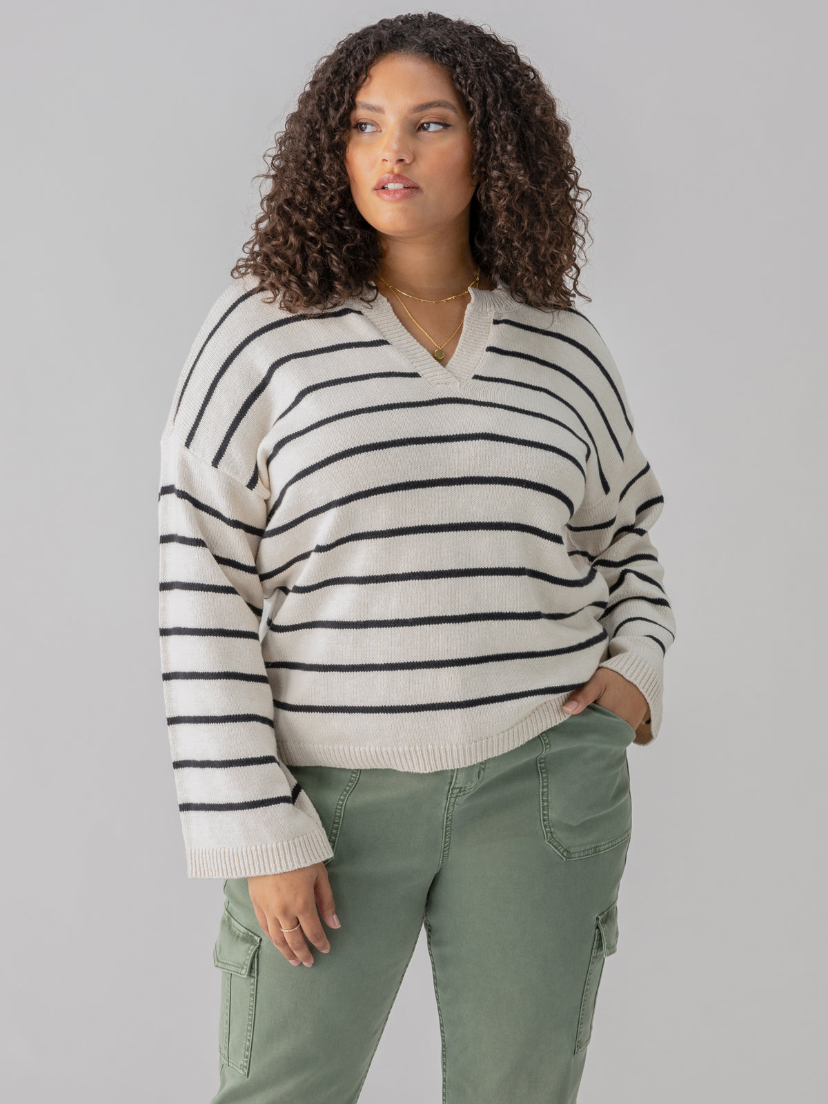 A person with curly hair is wearing the Chill Vibes Sweater Chalk Black Stripe from Sanctuary Clothing's Inclusive Collection. They have one hand in the pocket of their green cargo pants and are looking to the side with a neutral expression. The background is plain gray.