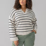 A person with curly hair is wearing the Chill Vibes Sweater Chalk Black Stripe from Sanctuary Clothing's Inclusive Collection. They have one hand in the pocket of their green cargo pants and are looking to the side with a neutral expression. The background is plain gray.