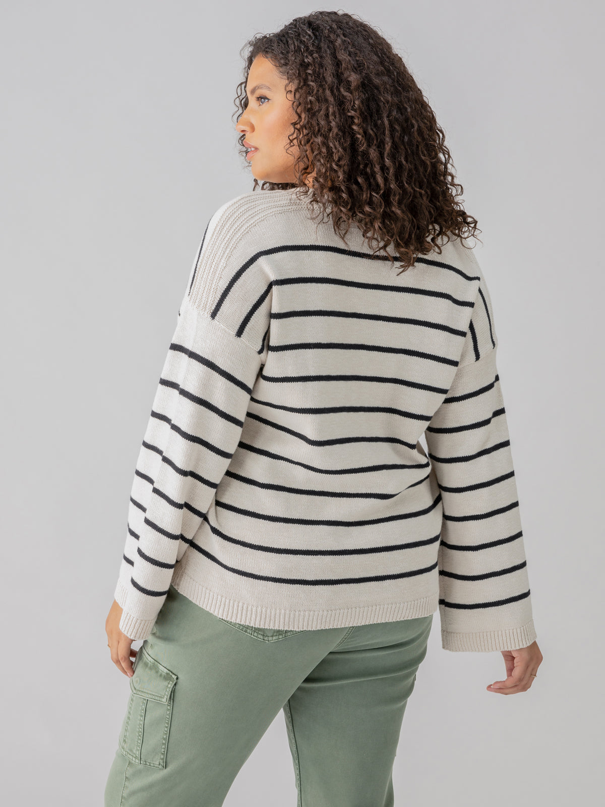 A person with curly hair stands with their back facing the camera, wearing the Chill Vibes Sweater Chalk Black Stripe from the Inclusive Collection by Sanctuary Clothing, paired with green cargo pants. The background is plain and gray.