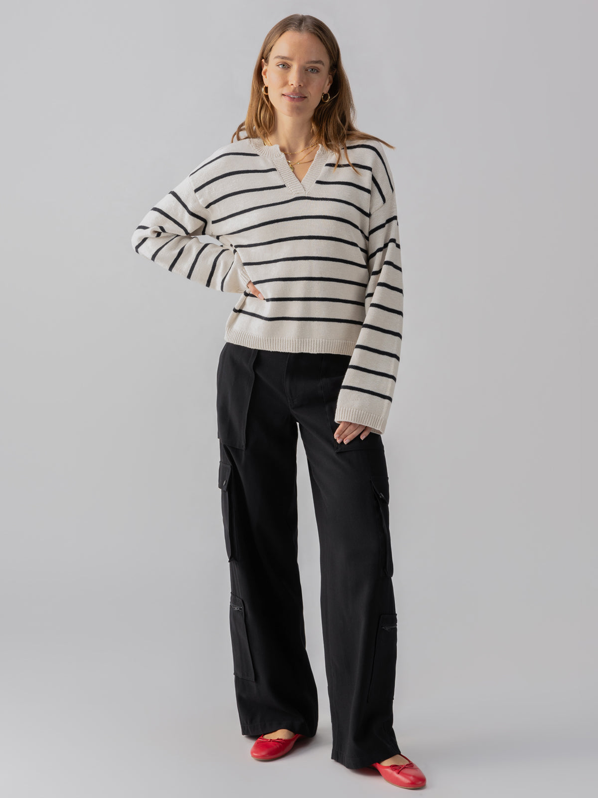 A person stands against a plain background wearing the Chill Vibes Sweater in Chalk Black Stripe by Sanctuary Clothing, black wide-leg pants with pockets, and red flat shoes. They have long hair and an arm resting on their hip.