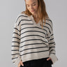 A person with long hair is wearing the Sanctuary Clothing Chill Vibes Sweater in Chalk Black Stripe and dark pants. They have their hands in their pockets, looking down with a slight smile against a plain, light-colored background.