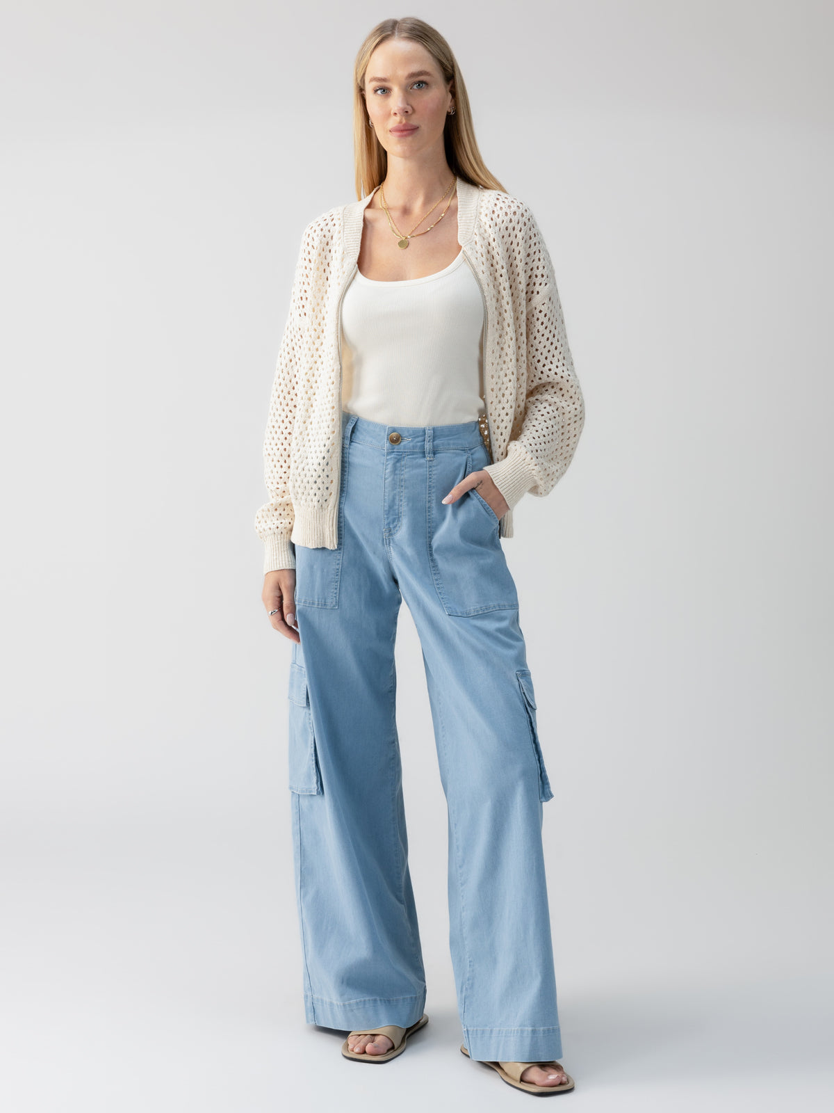 A person with long hair stands in a neutral pose against a plain background. They are wearing a white tank top, a cream-colored knit cardigan, the Sunset Denim Pant Pale Blue from Sanctuary Clothing, and sandals. One hand is in their pants pocket, and they wear a calm expression.