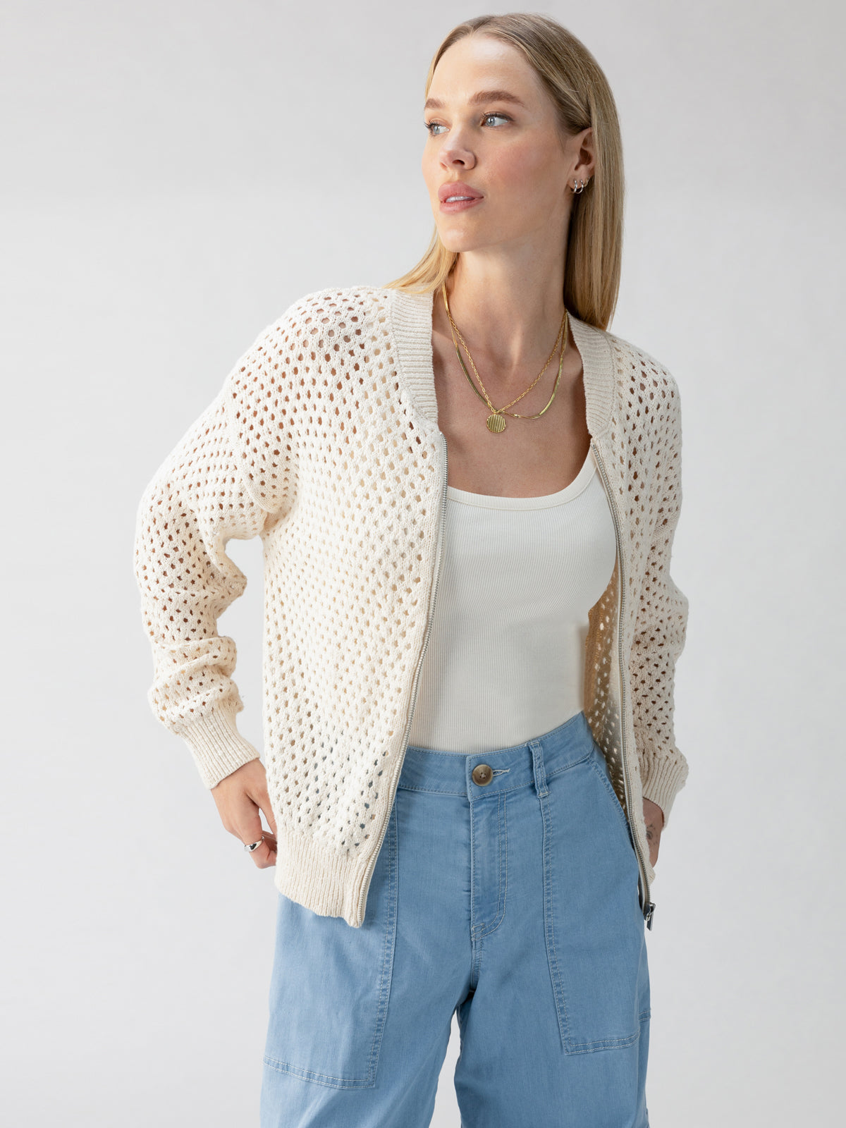 A person with light-colored hair wears the Stepping Out Bomber Eco Natural from Sanctuary Clothing over a white tank top and light blue pants. They are looking to the side with a neutral expression against a plain background. Multiple gold necklaces accessorize the outfit.