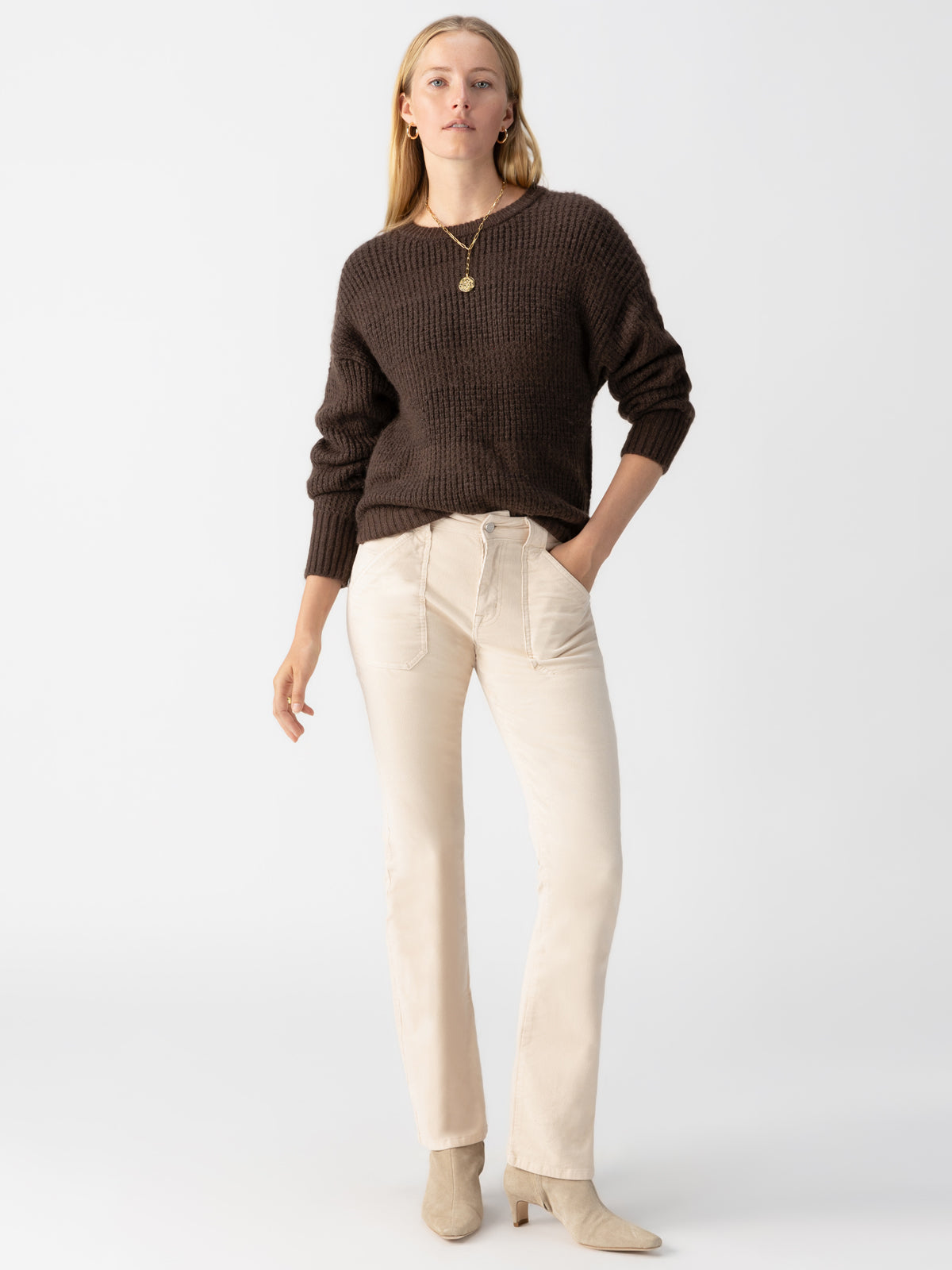 A woman standing against a plain background is modeling the Corduroy Hayden Bootcut Toasted Almond pants from Sanctuary Clothing, paired with a dark brown sweater and beige ankle boots. She accessorizes with a gold necklace, her left hand in her pocket and her right arm relaxed by her side.