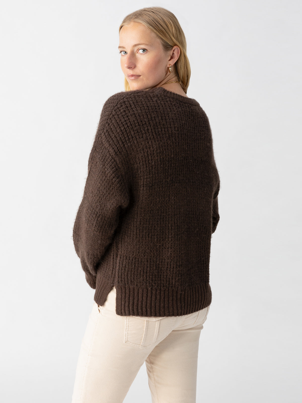 A person with blonde hair is standing against a light grey background, wearing the dark brown Snow Bunny Sweater Coffee by Sanctuary Clothing and beige pants. They are turned slightly to their right and looking over their left shoulder towards the camera.