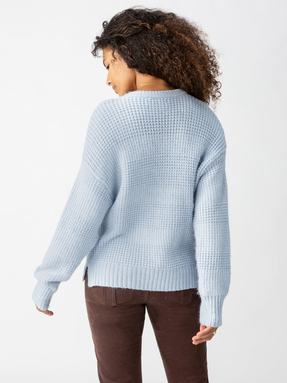 Person with curly hair wearing the Snow Bunny Sweater Sky by Sanctuary Clothing and brown pants, photographed from behind against a plain white background.