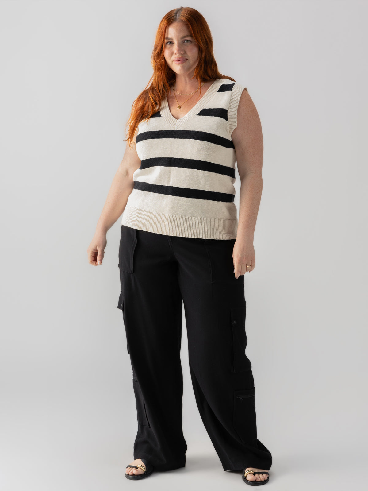 A woman with long red hair stands against a plain background, wearing the Sanctuary Clothing Sweater Shell Eco Natural Black Stripe from the Inclusive Collection and black cargo pants. She is looking at the camera with a neutral expression, her arms resting comfortably by her sides.