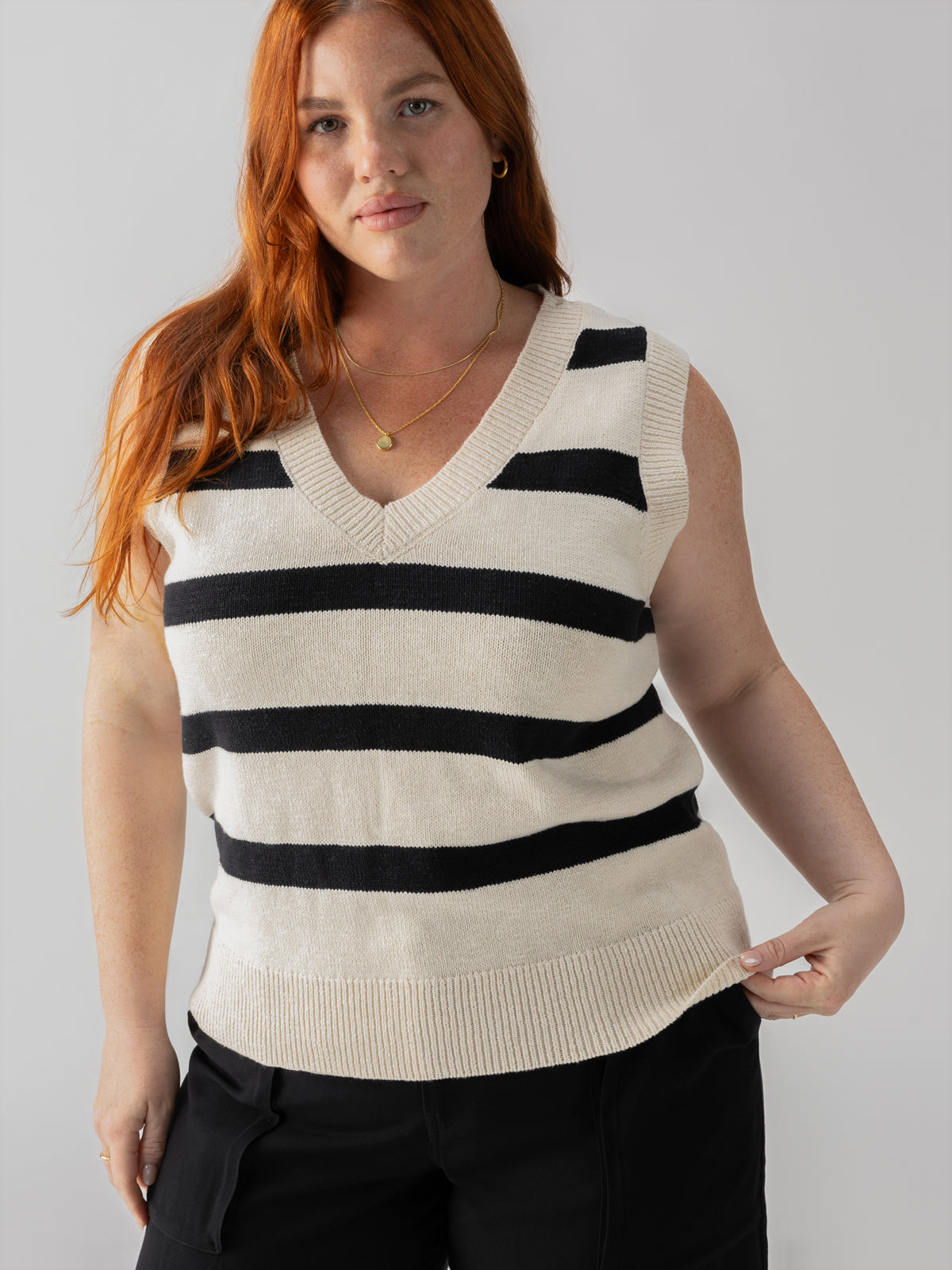 A woman with long red hair, wearing the Sanctuary Clothing Sweater Shell Eco Natural Black Stripe from the Inclusive Collection, is standing against a plain backdrop. She is also wearing black pants and a gold necklace. She has a relaxed expression and is holding the hem of her sweater.