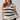 A woman with long red hair, wearing the Sanctuary Clothing Sweater Shell Eco Natural Black Stripe from the Inclusive Collection, is standing against a plain backdrop. She is also wearing black pants and a gold necklace. She has a relaxed expression and is holding the hem of her sweater.