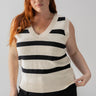 A woman with long red hair, wearing the Sanctuary Clothing Sweater Shell Eco Natural Black Stripe from the Inclusive Collection, is standing against a plain backdrop. She is also wearing black pants and a gold necklace. She has a relaxed expression and is holding the hem of her sweater.