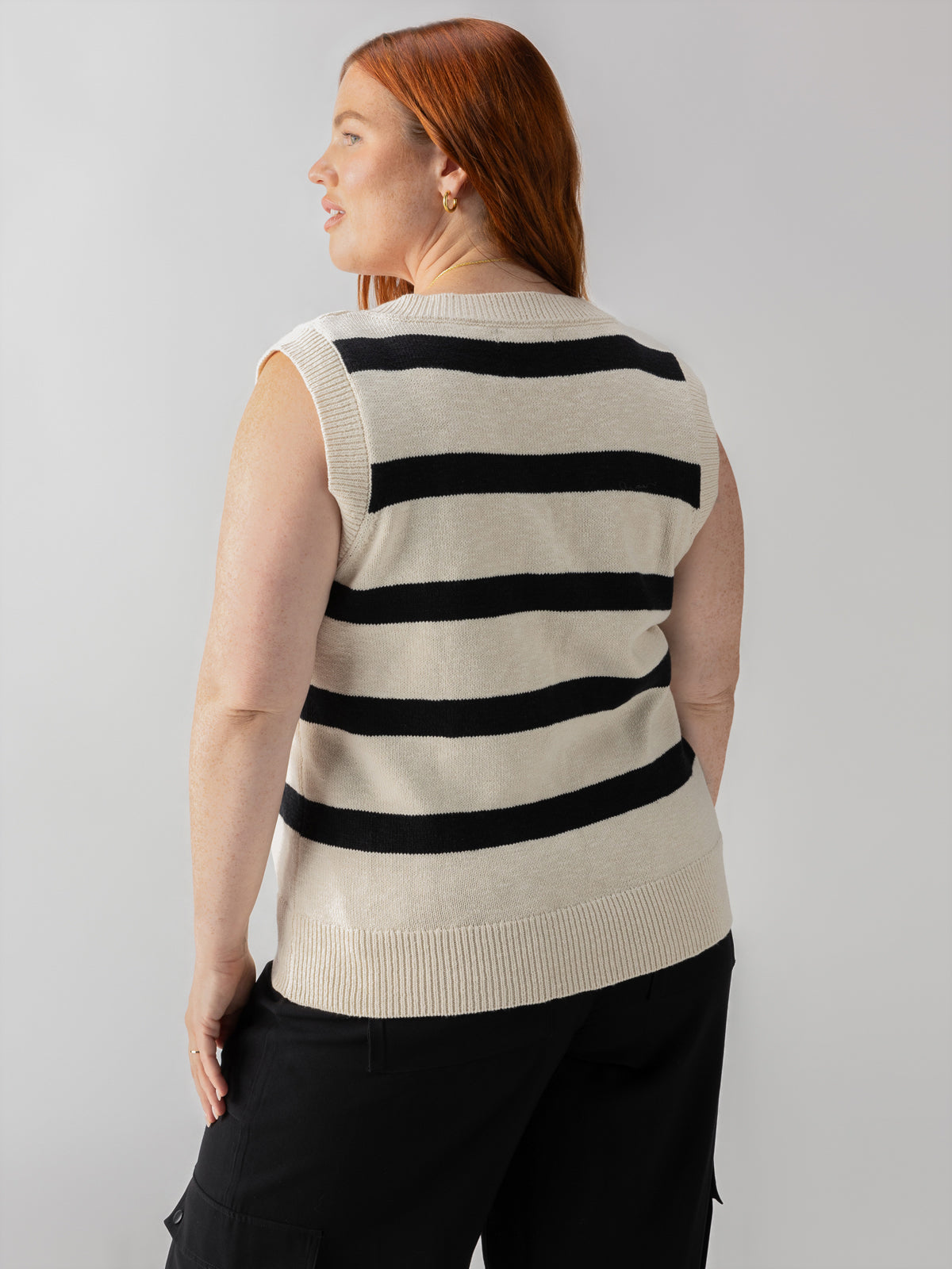 A person with long red hair is shown from the back, wearing a sleeveless beige sweater with black horizontal stripes. They are dressed in black pants and stand against a light gray background.