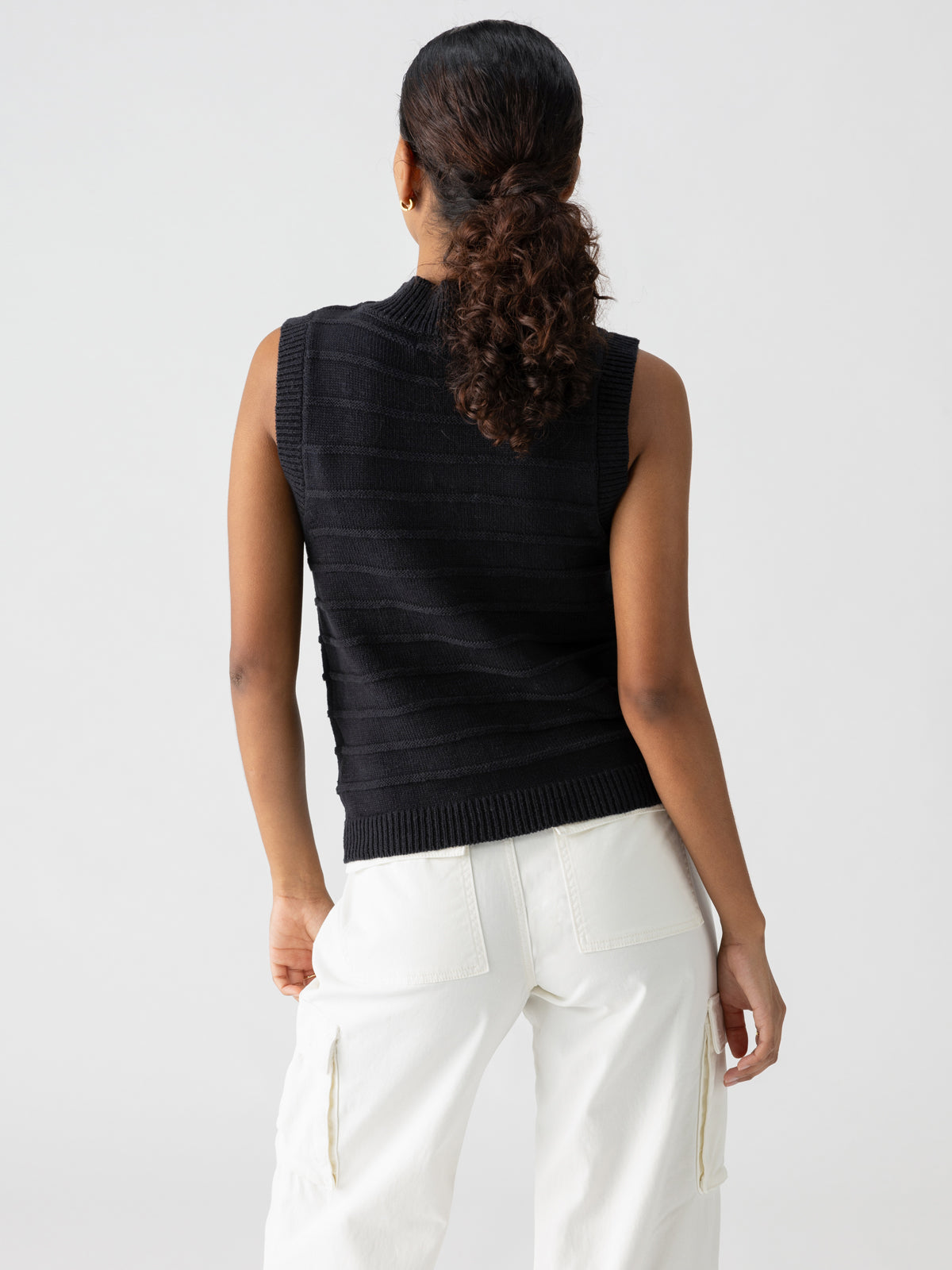 A person with curly hair in a ponytail is standing with their back to the camera. They are wearing a Sanctuary Clothing sleeveless top named Life Is Easy Shell Black and white pants with side pockets. The background is plain and light-colored.